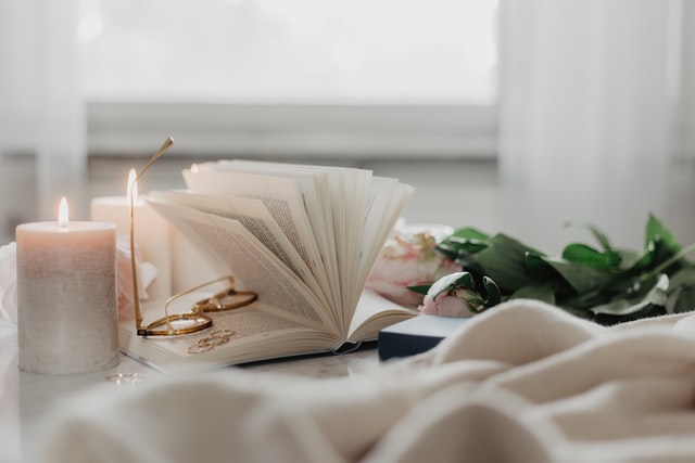 Mesa branca com livro aberto, óculos e velas brancas acesas.