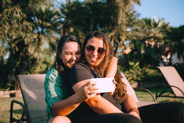 Mulheres brancas abraçadas, enquanto tiram fotos com um celular.