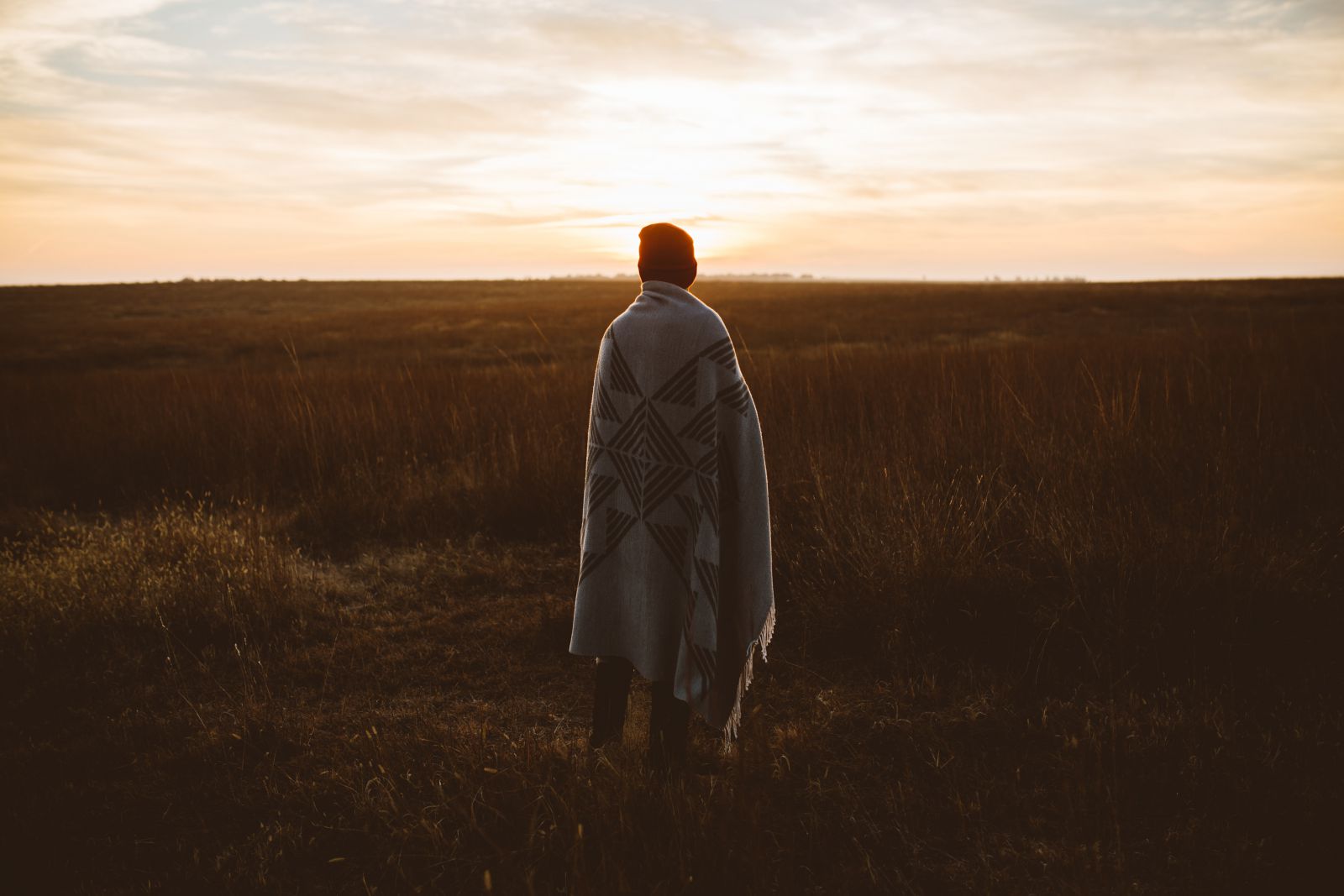 Pessoa em meio a um campo para o pôr do sol, usando touca e cobertor
