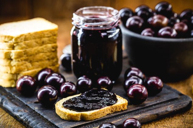 Pote com geleia de jabuticaba. À esquerda, fatias de pão empilhadas e algumas jabuticabas soltas. À direita, um recipiente cheio de jabuticabas. Na frente, uma fatia de pão coberta com geleia.