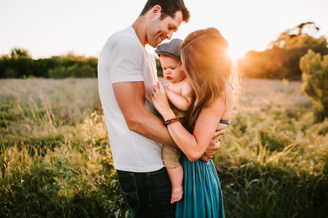 Homem e mulher brancos, segurando um bebê, no meio de um campo.