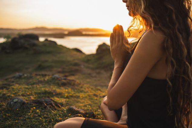 Mulher mostrada parcialmente, sentada em posição de meditação em um ambiente rochoso e gramado.