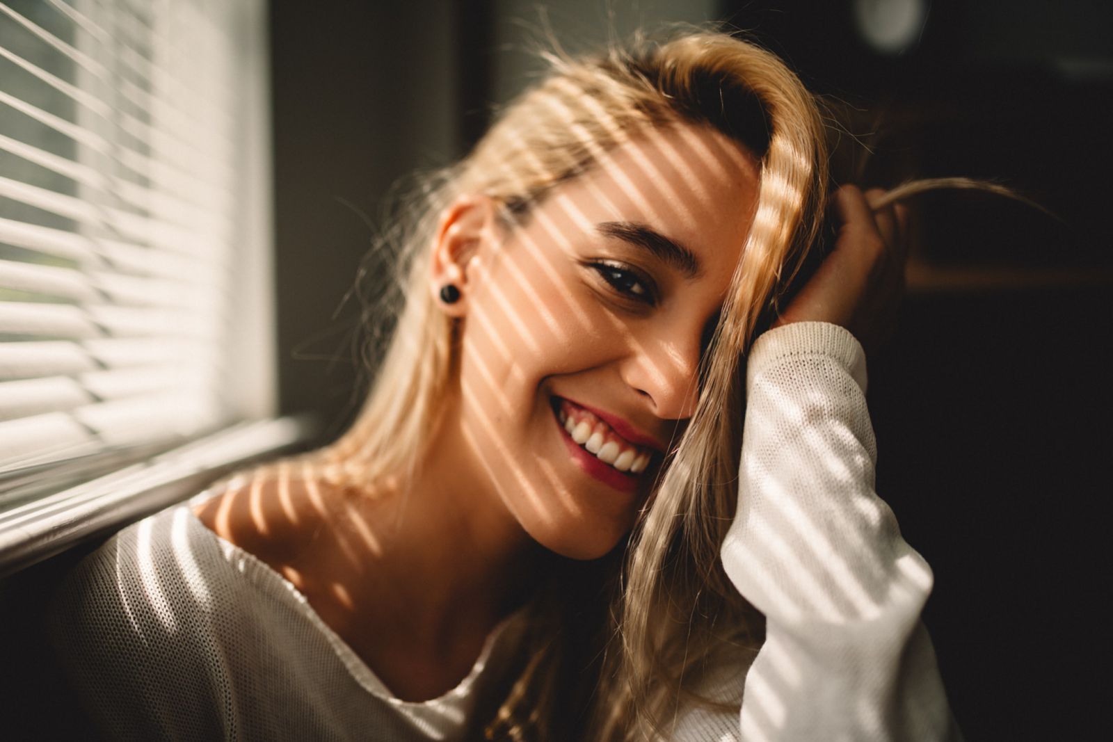 Mulher sorrindo segurando seu cabelo ao lado de uma janela