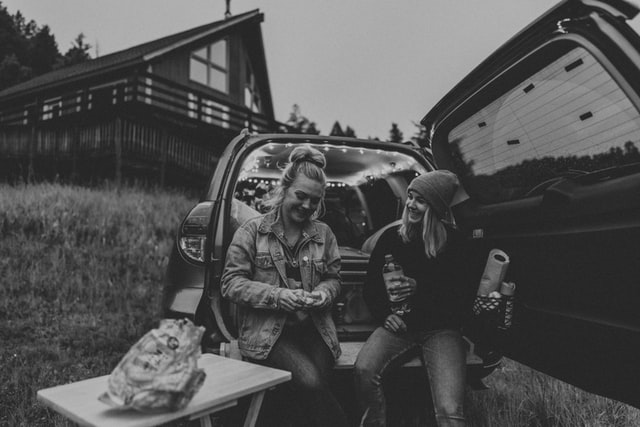 Mulheres brancas e loiras sentadas no porta-malas de um carro, com bebidas na mão.