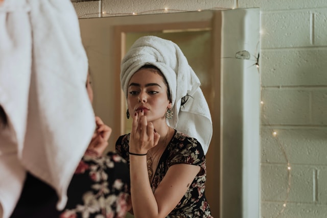 Mulher branca de toalha na cabeça enquanto passa batom.