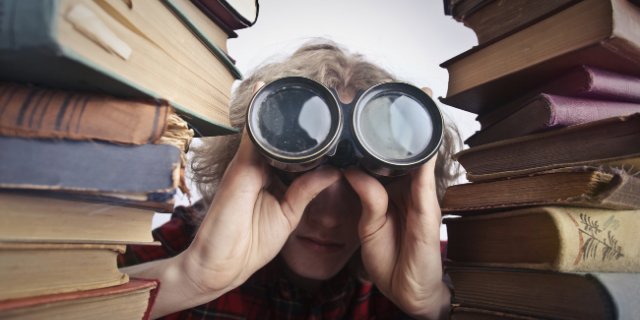Mulher com binóculos sobre os olhos. Ela está entre duas colunas de livros.