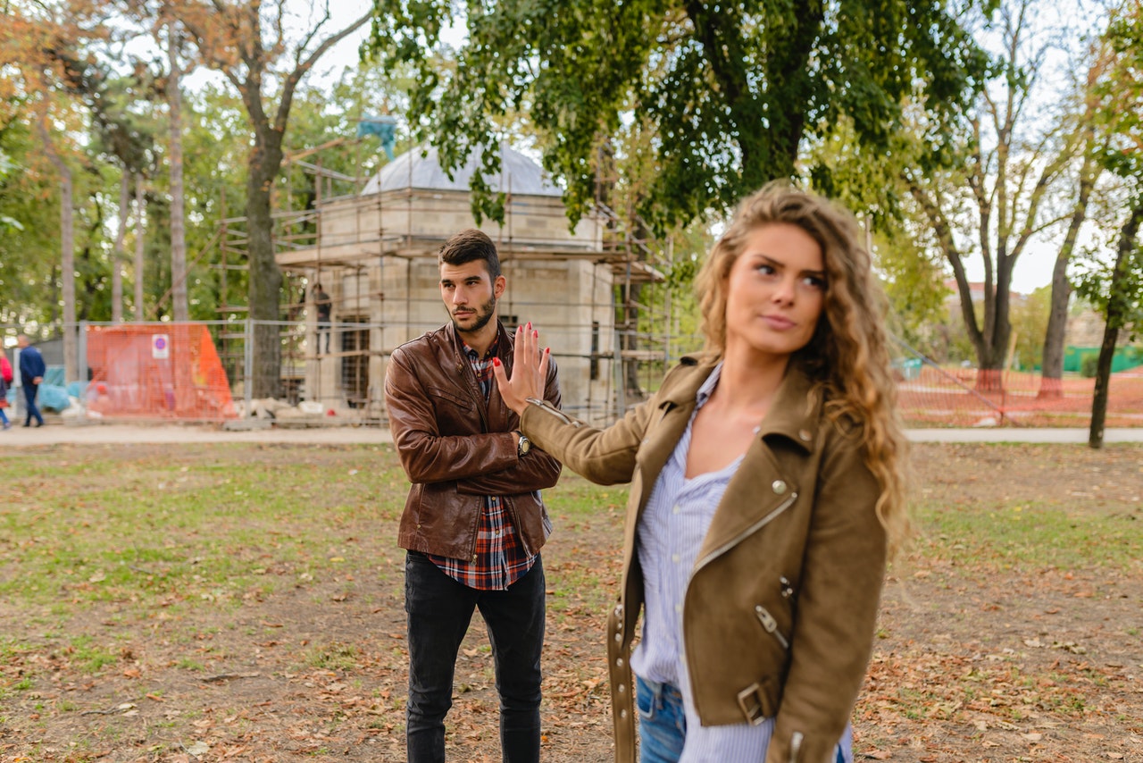 Mulher com o braço esticado para trás e a mão erguida, em direção a homem, para evitá-lo. O homem está de braços cruzados e com expressão de impaciente.