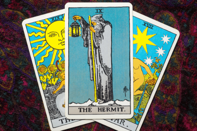A carta O Eremita do Tarot está em destaque sobre uma mesa. Ao lado, há outras duas cartas de Tarot.