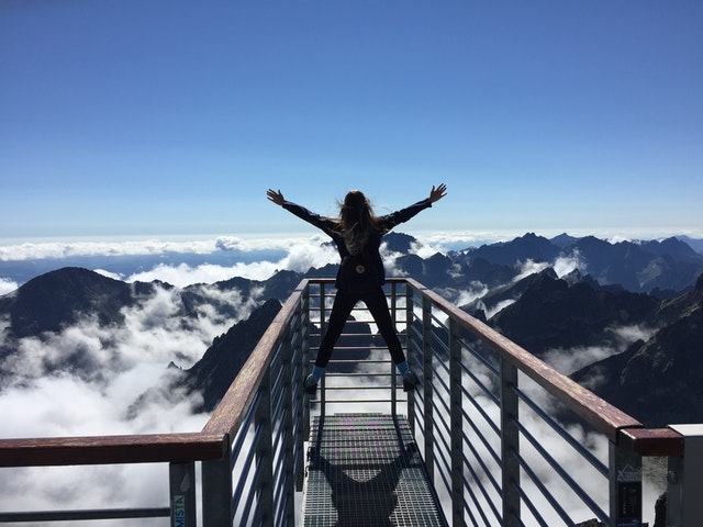 Mulher de costas em parapeito com nuvens e topo de montanhas ao fundo