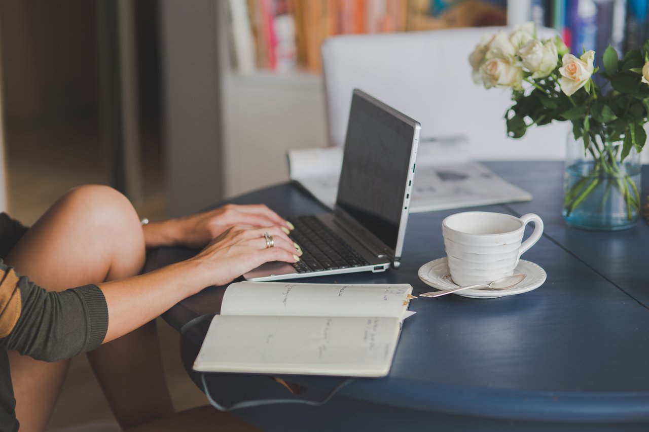 Mulher digitando em um notbook em cima de uma mesa com flores e uma xícara