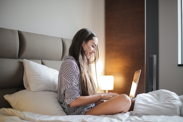 Mulher branca sentada numa cama com notebook no colo.