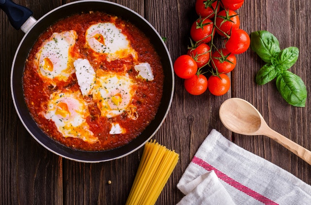 Mesa de madeira escura com panela, tomates, macarrão e folhas.