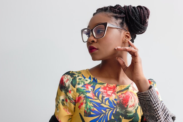 Mulher negra usando roupas coloridas e óculos, com mão levada ao rosto.
