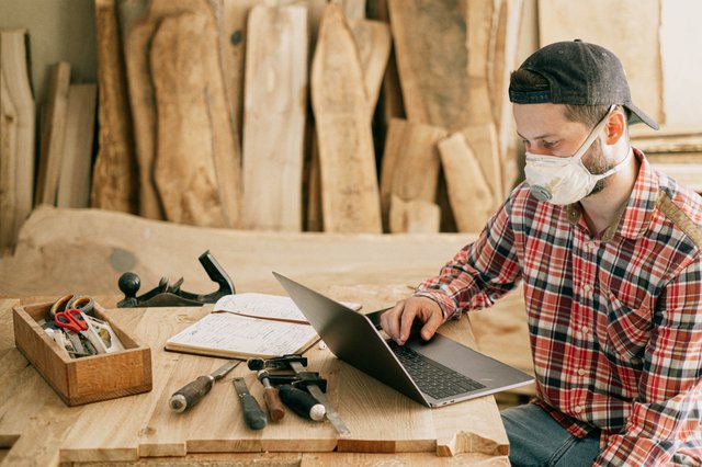 Homem branco em frente à notebook com madeiras e utensílios de marcenaria ao redor.