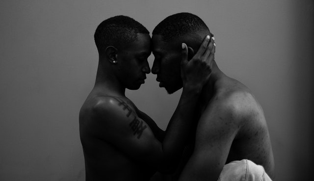 Dois homens negros com cabeças encostadas e olhos fechados. Mãos de um deles ao redor da cabeça do outro.