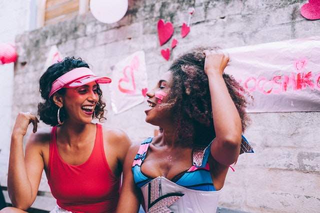 Mulher branca e mulher negra vestindo roupas e adereços coloridos.