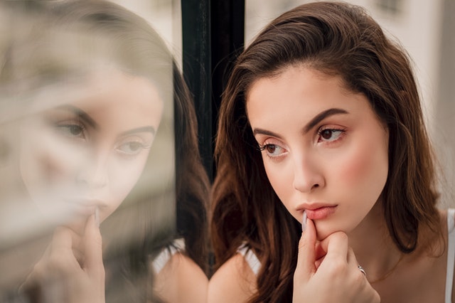 Mulher em frete a janela com reflexo aparecendo no vidro e expressão seria