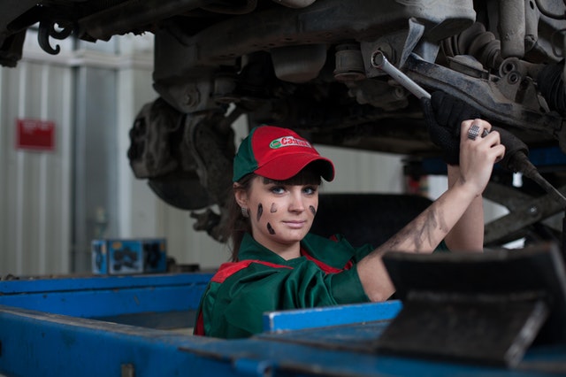 Mulher branca com roupas de mecânica enquanto arruma um automóvel.