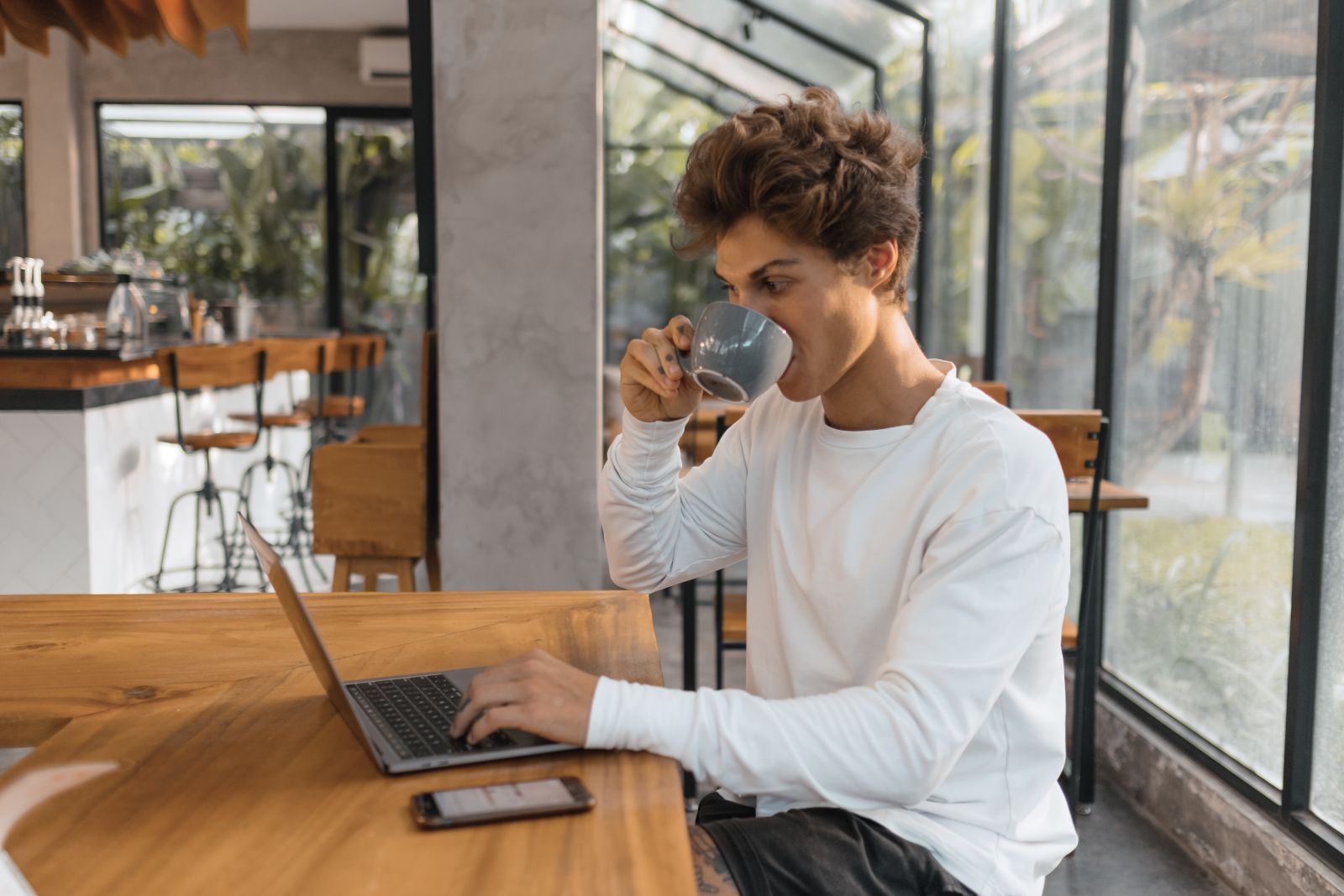 Homem sentado mexendo em seu computador enquanto está tomando algo em sua xícara