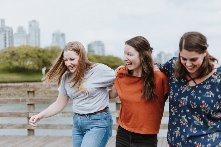 Três mulheres brancas com expressões sorridentes e abraçadas.