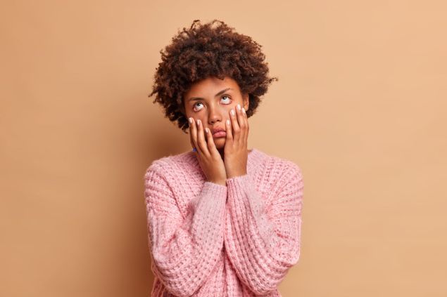 Mulher negra vestindo um casaco cor de rosa claro. Ela está revirando os olhos, com as mãos no rosto, demonstrando tédio.