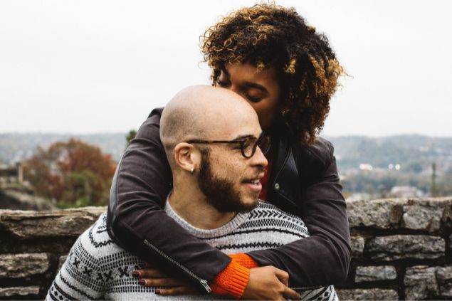 Casal formado por um homem e uma mulher. A mulher está atrás do homem, abraçando-o pelo pescoço e beijando sua cabeça.