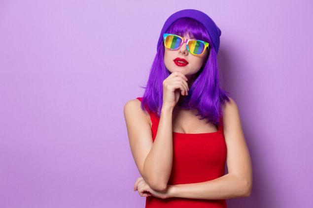 Mulher com cabelos na cor roxa, usando um óculos nas cores do arco-íris. Ela apoia uma mão levemente no queixo e olha para cima, sugerindo estar pensando.