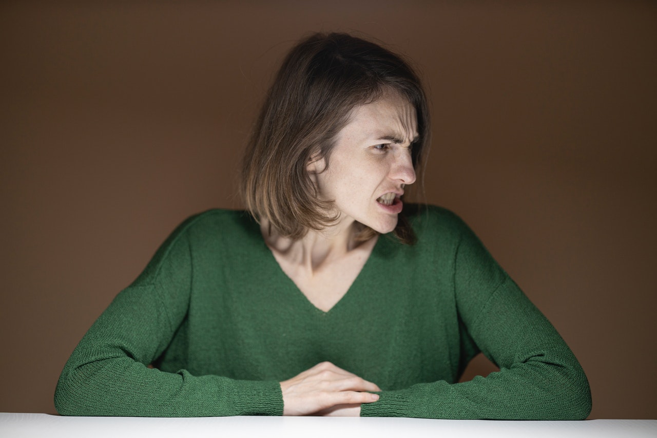 Mulher sentada, apoiando os braços em uma mesa, e olhando para o lado enquanto range os dentes.