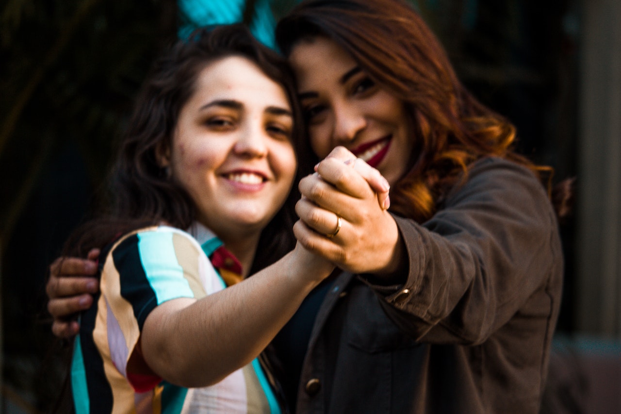 Duas mulheres com braços esticados e dedos entrelaçados, dançando, e olhando para a câmera.
